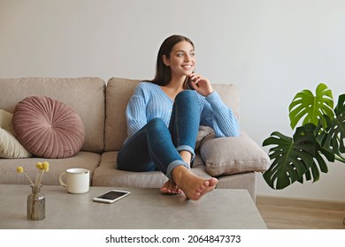 Kick Back And Relax Concept. Young Beautiful Brunette Woman With Blissful Facial Expression Alone On The Couch With Her Bare Feet On Coffee Table. Portrait Of Relaxed Female Resting At Home.