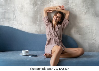 Kick Back And Relax Concept. Young Woman With Blissful Facial Expression Sitting On Couch At Home Next To Coffee Mug And Cellphone, Stretching Hands On Sunday Morning With Closed Eyes