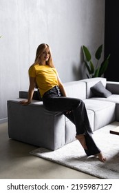 Kick Back And Relax Concept. Young Beautiful Brunette Woman With Blissful Facial Expression Alone On The Couch With Her Bare Feet On Coffee Table. Portrait Of Relaxed Female Resting At Home.