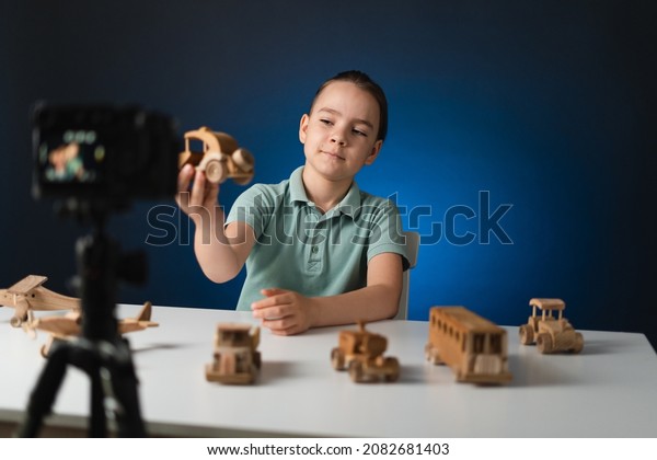 Kid blogger filming\
video on camera indoors with blue light in background. Online\
reviewing. Promoting eco wooden toys. Kid recording his gameplay\
for vlog.