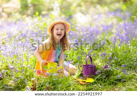 Kid in bluebell woodland. Child with flowers, garden tools and wheelbarrow. Girl gardening. Children play outdoor in bluebells, work, plant and water blue bell flower bed. Family fun in summer forest.