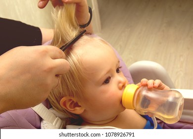 Kid boy with blond hairs and with bottle getting his first haircut. Close up of hands of hairdresser. 