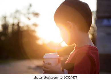 Kid Boy Drinks Cocoa Or Soda In A Cup With Tube On Sunset Background. Silhouette Of A Drinking Boy At Sunset.