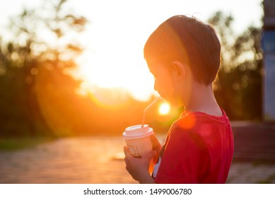 Kid Boy Drinks Cocoa Or Soda In A Cup With Tube On Sunset Background. Silhouette Of A Drinking Boy At Sunset