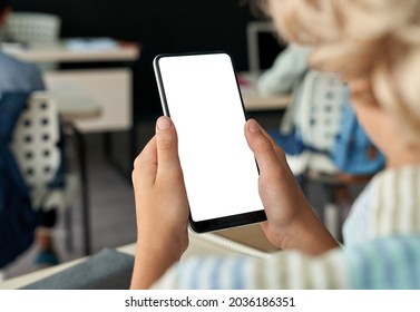 Kid Boy Junior School Student Using Smartphone Apps Holding Mobile Phone With White Blank Empty Mockup Screen In Classroom. Online Education, Mobile Application Classes Ads Concept. Over Shoulder View