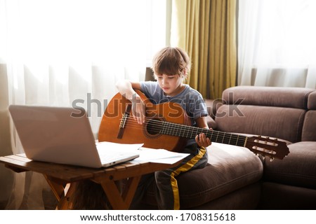 Kid  boy playing  guitar and watching  online lessons  on laptop while practicing at home.  Stay home. quarantine. Online training, online classes.
