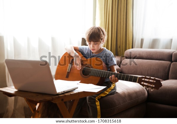 Kid  boy playing  guitar and watching  online\
lessons  on laptop while practicing at home.  Stay home.\
quarantine. Online training, online\
classes.\
