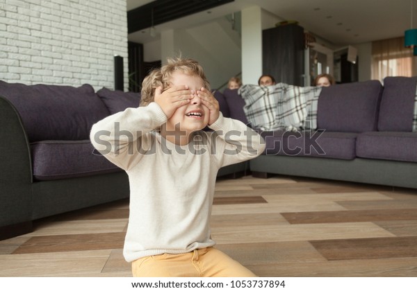Kid boy playing hide and seek game at home, child\
closing eyes with hands counting while parents and sister hide\
behind sofa in living room peeking out, happy family having fun\
with children concept