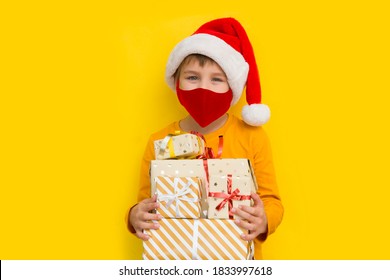 Kid Boy  In A Santa Hat Wearing Medical Face Mask  With Christmas Gift Boxes Against Yellow Background. Christmas Shopping.  Copy Space. New Normal.