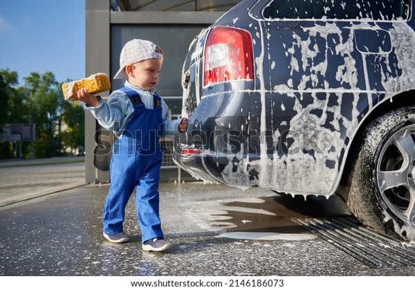 Kid boy studying\
work at car wash. Toddler child, holding yellow foam sponge in his\
right hand, examining soap car and preparing to start cleaning the\
back part of transport.