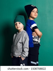 Kid Boy And Teenager In Modern Sportswear Knitted Hats, Pants, Hoody And T-shirt Cool Posing Back To Back With Hands In Their Pockets And Crossed On Green Background