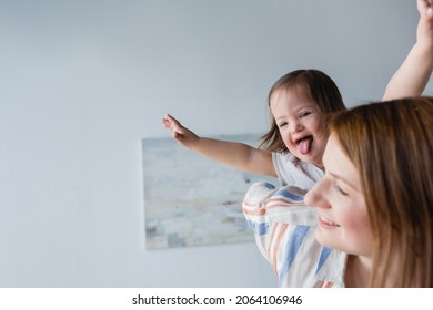 Kid with down syndrome sticking out tongue near blurred mother at home - Shutterstock ID 2064106946