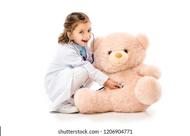 kid dressed in doctors white coat with stethoscope playing with teddy bear isolated on white
