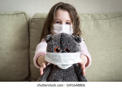 Kid In Face Mask For Protection To Corona Virus Playing At Home, Funny Little Girl And Toy In Room During COVID-19 Pandemic. Child Staying In House Due To Coronavirus. Safety And Quarantine Concept.