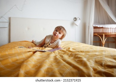 kid flips through a book on bed in bedroom, girl Toddler in overalls lies on bed with book