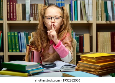 Kid Girl Asks To Be Quiet In Library, School Child Sits Alone At Table With Books, Holding One Finger On Mouth, Keep Silence Concept