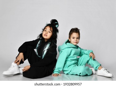 Kid Girl In Black Hoodie And Pants And Kid Boy In Pastel Modern Green, Mint Color Sports Suit Are Sitting Back To Back On Floor Over White Background. Friendship And Funky Time