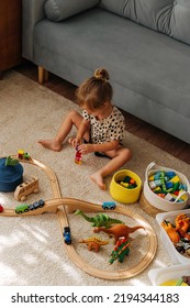 Kid Girl Builds A Toy Town And  Wooden Railway. A Little Girl Sitting On Floor Plays With Toy Train Locomotive,  Wooden Blocks And Toy Animals. Educational Game For Toddler In Modern Nursery.