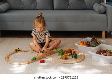 Kid Girl Builds A Toy Town And  Wooden Railway. A Little Girl Sitting On Floor Plays With Toy Train Locomotive,  Wooden Blocks And Toy Animals. Educational Game For Toddler In Modern Nursery.