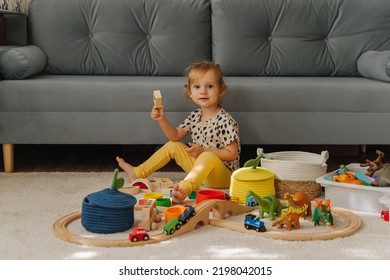 Kid Girl Builds A Toy Town And  Wooden Railway. A Little Girl Sitting On Floor Plays With Toy Train Locomotive,  Wooden Blocks And Toy Animals. Educational Game For Toddler In Modern Nursery.