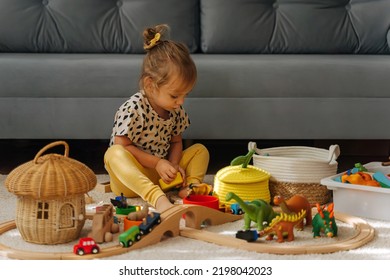 Kid Girl Builds A Toy Town And  Wooden Railway. A Little Girl Sitting On Floor Plays With Toy Train Locomotive,  Wooden Blocks And Toy Animals. Educational Game For Toddler In Modern Nursery.