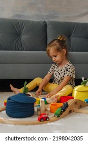 Kid Girl Builds A Toy Town And  Wooden Railway. A Little Girl Sitting On Floor Plays With Toy Train Locomotive,  Wooden Blocks And Toy Animals. Educational Game For Toddler In Modern Nursery.