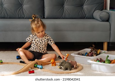 Kid Girl Builds A Toy Town And  Wooden Railway. A Little Girl Sitting On Floor Plays With Toy Train Locomotive,  Wooden Blocks And Toy Animals. Educational Game For Toddler In Modern Nursery.