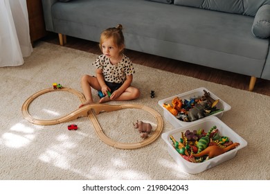 Kid Girl Builds A Toy Town And  Wooden Railway. A Little Girl Sitting On Floor Plays With Toy Train Locomotive,  Wooden Blocks And Toy Animals. Educational Game For Toddler In Modern Nursery.