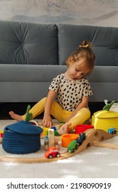 Kid Girl Builds A Toy Town And  Wooden Railway. A Little Girl Sitting On Floor Plays With Toy Train Locomotive,  Wooden Blocks And Toy Animals. Educational Game For Toddler In Modern Nursery.