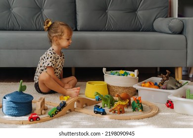 Kid Girl Builds A Toy Town And  Wooden Railway. A Little Girl Sitting On Floor Plays With Toy Train Locomotive,  Wooden Blocks And Toy Animals. Educational Game For Toddler In Modern Nursery.