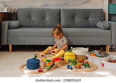 Kid Girl Builds A Toy Town And  Wooden Railway. A Little Girl Sitting On Floor Plays With Toy Train Locomotive,  Wooden Blocks And Toy Animals. Educational Game For Toddler In Modern Nursery.