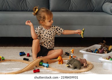 Kid Girl Builds A Toy Town And  Wooden Railway. A Little Girl Sitting On Floor Plays With Toy Train Locomotive,  Wooden Blocks And Toy Animals. Educational Game For Toddler In Modern Nursery.