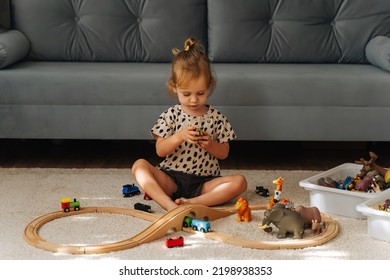 Kid Girl Builds A Toy Town And  Wooden Railway. A Little Girl Sitting On Floor Plays With Toy Train Locomotive,  Wooden Blocks And Toy Animals. Educational Game For Toddler In Modern Nursery.