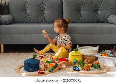 Kid Girl Builds A Toy Town And  Wooden Railway. A Little Girl Sitting On Floor Plays With Toy Train Locomotive,  Wooden Blocks And Toy Animals. Educational Game For Toddler In Modern Nursery.