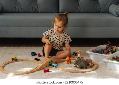 Kid Girl Builds A Toy Town And  Wooden Railway. A Little Girl Sitting On Floor Plays With Toy Train Locomotive,  Wooden Blocks And Toy Animals. Educational Game For Toddler In Modern Nursery.