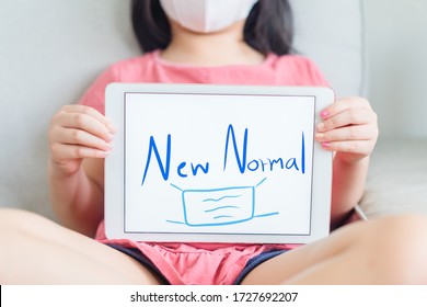 Kid Girl Holding Tablet And Writing NEW NORMAL Word With Face Mask Picture Showing At Home.New Normal Post Covid-19 Crisis Change Human Behavior.Concept For Stay Home,Wellness Health, Digital Takeover