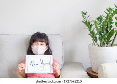 Kid Girl Holding Tablet And Writing NEW NORMAL Word With Face Mask Picture Showing At Home.New Normal Post Covid-19 Crisis Change Human Behavior.Concept For Stay Home,Wellness Health, Digital Takeover