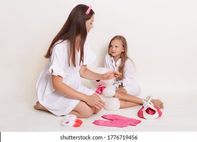 Kid Girl And Pregnant Mother Sitting On Floor Surrounded With Child's Clothing And Soft Baby, Expectant Mother Wearing White Dress And Rose Hair Band Sitting On Floor With Elder Child.