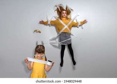 A Kid Girl In A Yellow Dress Smiles Sinisterly And Holds Duct Tape In Her Hands. Young Mom  And Banana Glued To The Wall With Adhesive Tape. The Child Casts The Shadow Of The Devil With Horns