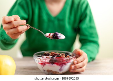 A Kid With Green Shirt Is Eating Fresh Homemade Glass Bowl Of Creamy Yogurt Parfait With Berries, Muesli And Seeds In It On Wooden Table Closeup Photo Shows Hand Holding A Spoon With An Apple On Side.