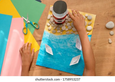 Kid Hands Make A Lighthouse For School Project At Home. Top Down View To Wooden Table With Colored Paper And Other Supplies