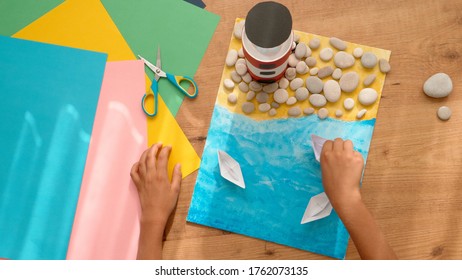 Kid Hands Make A Lighthouse For School Project At Home. Top Down View To Wooden Table With Colored Paper And Other Supplies