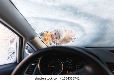 The Kid Helps And Scraping Snow And Ice From Car Window. Girl Is Cleaning Car From Ice. View From Inside Of Car.