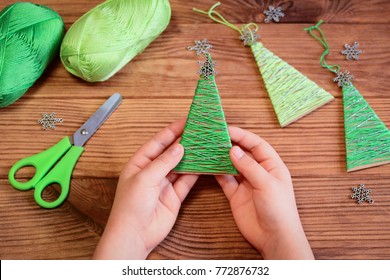 Kid Is Holding A DIY Yarn Christmas Tree Decoration In Hands. Kid Is Showing A DIY Yarn Christmas Tree Decoration. Merry Christmas Tree Project For Kids. Scissors, Green Cotton Yarn On A Wooden Table