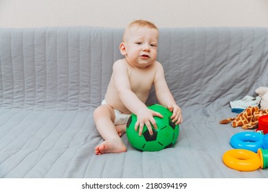 The Kid Is Holding A Soccer Ball, Sitting On The Couch Barefoot And Looking Away. Adorable Baby Is Playing At Home On The Couch. The Concept Of Holidays, Weekends And Childhood