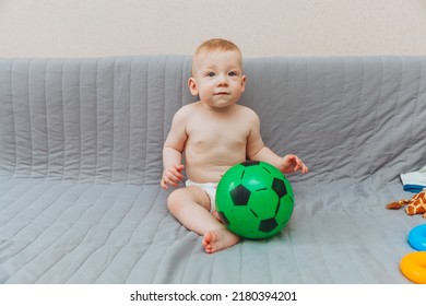 The Kid Is Holding A Soccer Ball, Sitting On The Couch Barefoot And Looking Away. Adorable Baby Is Playing At Home On The Couch. The Concept Of Holidays, Weekends And Childhood