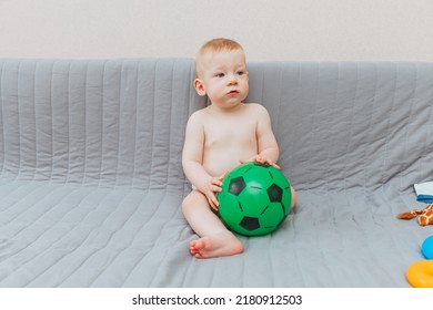 The Kid Is Holding A Soccer Ball, Sitting On The Couch Barefoot And Looking Away. Adorable Baby Is Playing At Home On The Couch. The Concept Of Holidays, Weekends And Childhood