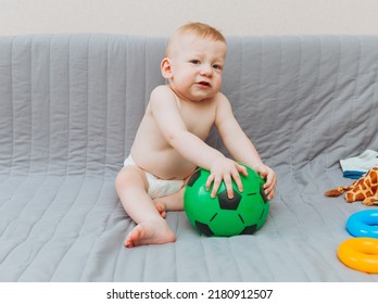 The Kid Is Holding A Soccer Ball, Sitting On The Couch Barefoot And Looking Away. Adorable Baby Is Playing At Home On The Couch. The Concept Of Holidays, Weekends And Childhood