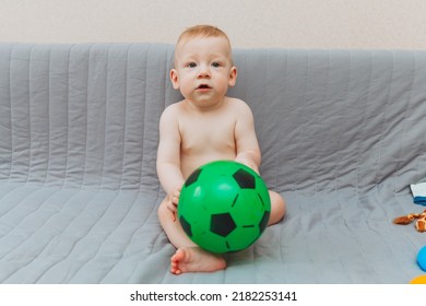 The Kid Is Holding A Soccer Ball, Sitting On The Couch Barefoot And Looking Away. Adorable Baby Is Playing At Home On The Couch. The Concept Of Holidays, Weekends And Childhood