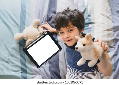 Kid laying in bed with dog toy and looking up at camera,Cute child boy lying in bed with tablet,Top view Children playing alone in bedroom with mock up of digital tablet.New normal lifestyle  - Powered by Shutterstock